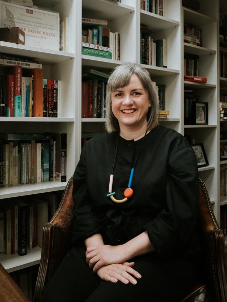 An image of Amber Massie-Blomfield, a woman dressed in a black with a bright coloured necklace on. She has grey hair cut in to a bob, with a fringe slightly pulled back from her face. She is sitting in a chair in front of a series of bookshelves filled with books.