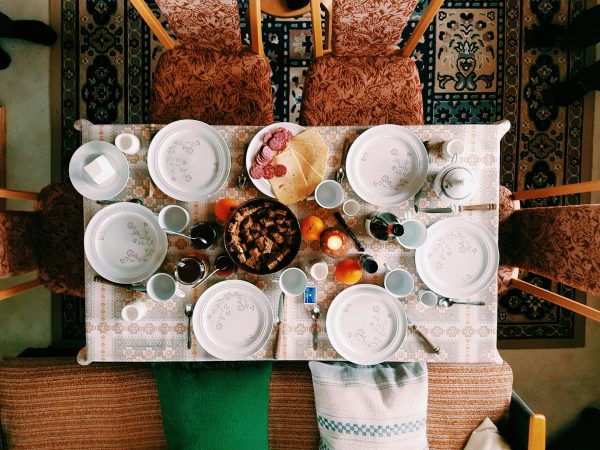 An overhead view of a table laid for 6 people. The plates are empty but there are dishes in the centre of the table waiting to be served, plus drinks and snacks. The table is surrounded by patterned chairs and several cushions with a bright patterned rug underneath the table.
