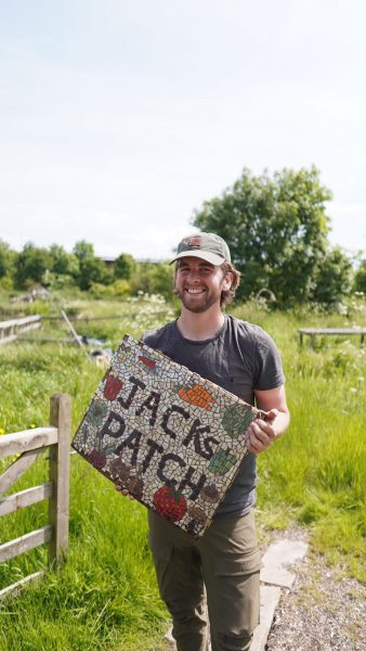 A bearded man in a baseball cap, standing in a sunny green field, holding a sign that says 'Jack's Patch'.